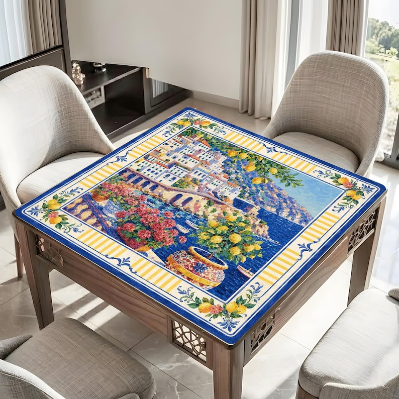 Puede incluir: Una mesa cuadrada de madera con una encimera de mosaico detallada y colorida. El mosaico representa una escena costera italiana con edificios, flores y un mar azul. La mesa est&aacute; rodeada de tres sillas.