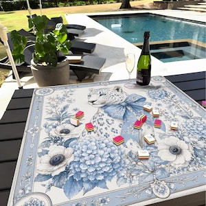 May include: A square game mat featuring a central illustration of a leopard wearing a large blue bow, surrounded by blue and white floral patterns including hydrangeas and anemones. Several small rectangular game tiles are scattered across the surface.