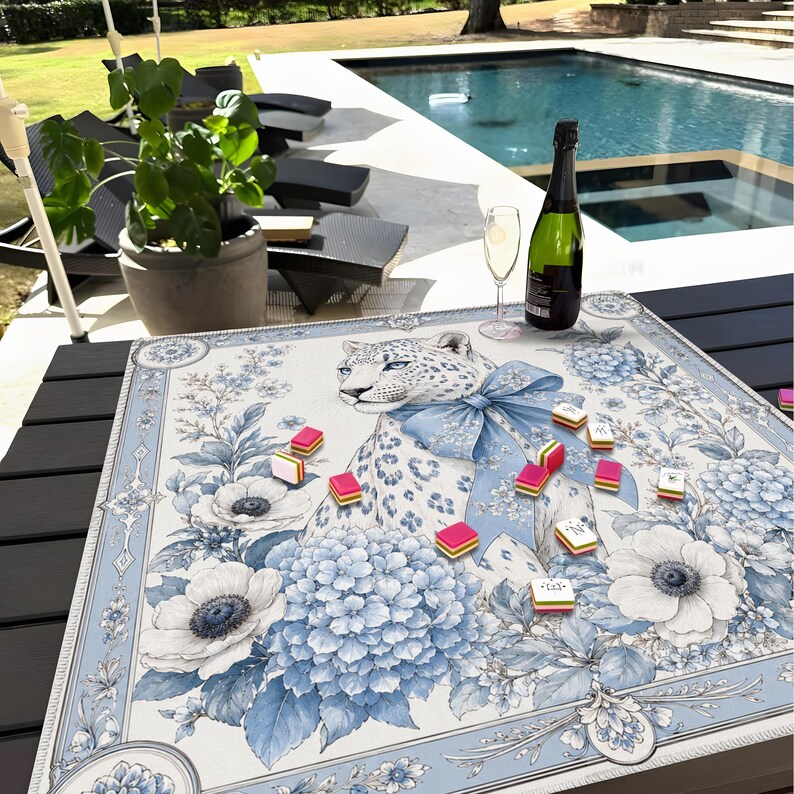 May include: A square game mat featuring a central illustration of a leopard wearing a large blue bow, surrounded by blue and white floral patterns including hydrangeas and anemones. Several small rectangular game tiles are scattered across the surface.