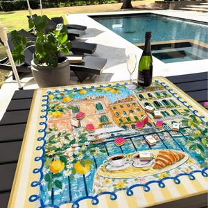May include: A vibrant table mat with a European town scene illustration, including buildings, lemons, and flowers. A bottle of champagne and a glass are on the table, alongside a game and a plate with a croissant and coffee.