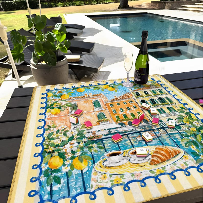 May include: A vibrant table mat with a European town scene illustration, including buildings, lemons, and flowers. A bottle of champagne and a glass are on the table, alongside a game and a plate with a croissant and coffee.