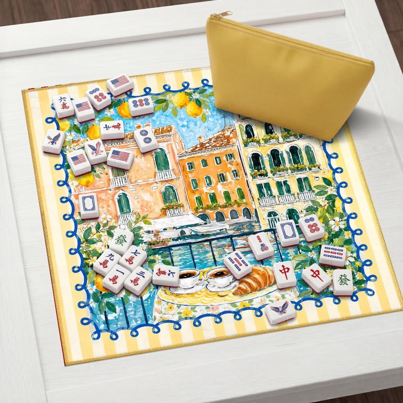May include: A yellow zipped pouch sits on a decorative mat depicting a European city scene. White Mahjong tiles with symbols and American flag designs are scattered around. The mat has a yellow and white striped border. The scene is reminiscent of Venice.