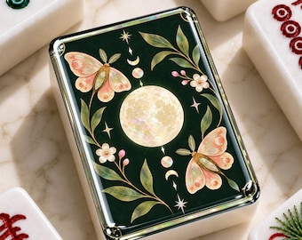 Puede incluir: Una ficha de mahjong rectangular con una superficie verde oscuro que presenta una luna llena central, dos polillas rosas, pequeñas flores blancas, hojas verdes y una secuencia vertical de fases lunares, rodeada por otras cuatro fichas blancas.