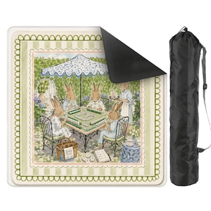 May include: A square outdoor blanket featuring a watercolor illustration of four rabbits playing mahjong under a blue and white umbrella, with a light green striped border and a scalloped edge. Includes a black cylindrical carrying bag with a strap.