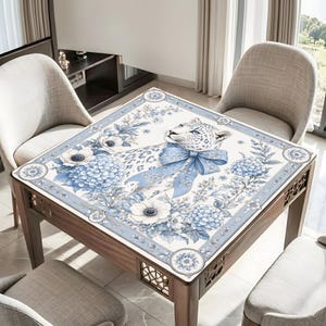 May include: A square table cover featuring a white leopard with blue spots, adorned with a large blue bow around its neck. The design includes blue and white floral patterns, including hydrangeas and anemones, set within a decorative border.