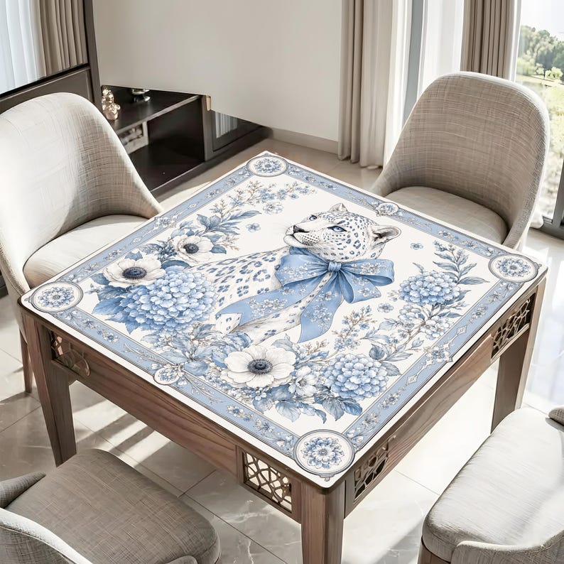 May include: A square table cover featuring a white leopard with blue spots, adorned with a large blue bow around its neck. The design includes blue and white floral patterns, including hydrangeas and anemones, set within a decorative border.