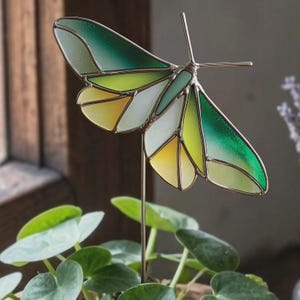 May include: A stained glass butterfly garden stake with a metal stem. The butterfly's wings feature shades of green, yellow, and white, with a brown metal outline. The stake is placed in a pot with green leafy plants.