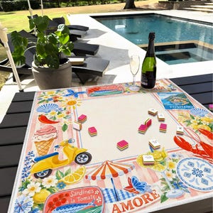 May include: A decorative table mat with a colorful Italian theme. The mat features illustrations of a yellow scooter, ice cream, lemons, and the word "AMORE". A bottle of champagne and a glass of champagne are on the table.