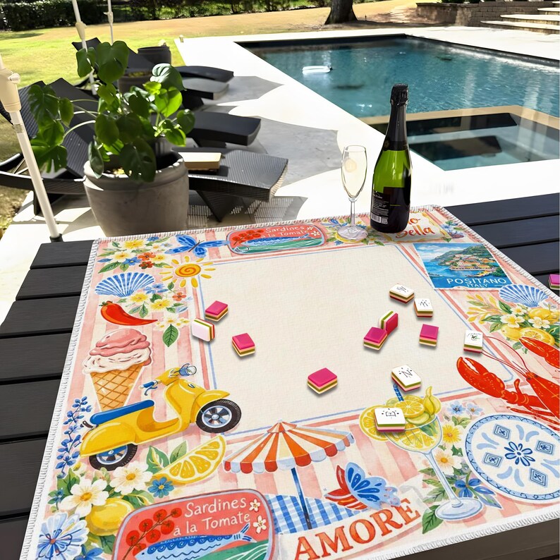 May include: A decorative table mat with a colorful Italian theme. The mat features illustrations of a yellow scooter, ice cream, lemons, and the word "AMORE". A bottle of champagne and a glass of champagne are on the table.