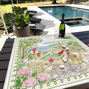 May include: A colorful Mahjong game set up on a decorative mat with a scenic illustration. The mat features a garden scene with pink flowers, a table, and a dog. A bottle of champagne and a glass are also present.