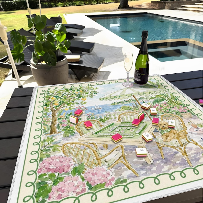 May include: A colorful Mahjong game set up on a decorative mat with a scenic illustration. The mat features a garden scene with pink flowers, a table, and a dog. A bottle of champagne and a glass are also present.