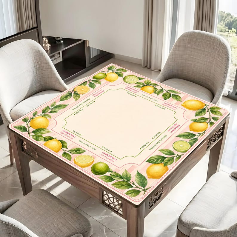 May include: A square table mat with a lemon and lime design. The mat features a border of lemons and green leaves, with a pink and white striped pattern. The center is a light beige color, with text around the edges.