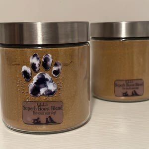 May include: Two glass jars with silver lids filled with a tan powder. One jar has a paw print sticker with a black and white design. The sticker reads "C.L.L.'s Superb Boost Blend For you & your dog!"