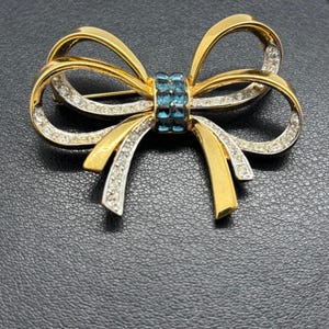 May include: A gold-tone bow brooch featuring a central cluster of square-cut blue crystals. The ribbon loops are accented with rows of clear pavé-set rhinestones along the edges, creating a layered metallic and sparkling appearance in this accessory.