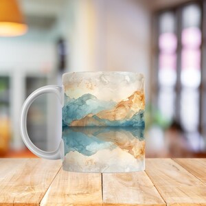 Può includere: Tazza da caffè in ceramica bianca con un design paesaggistico ad acquerello. Il design include immagini di montagne blu, beige e bianche riflesse nell'acqua. La tazza ha un manico curvo ed è appoggiata su una superficie di legno.