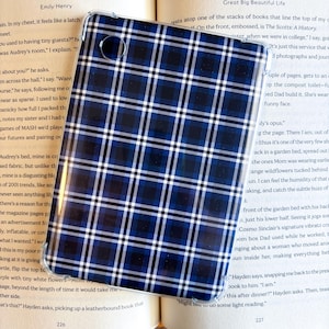 May include: A clear phone case with a blue, black, and white plaid pattern. The case is lying on top of an open book. The book's pages are visible, with text and page numbers.