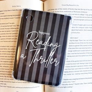 May include: A clear phone case with a black and gray striped design, overlaying an open book. The case features the words "happiest: Reading a Thriller" in white script.