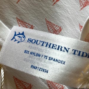 May include: Close-up of a white fabric with a repeating pattern of red sailboat illustrations. A white tag with blue text reads "SOUTHERN TIDE" and "93% NYLON / 7% SPANDEX". The tag also features a fish logo.
