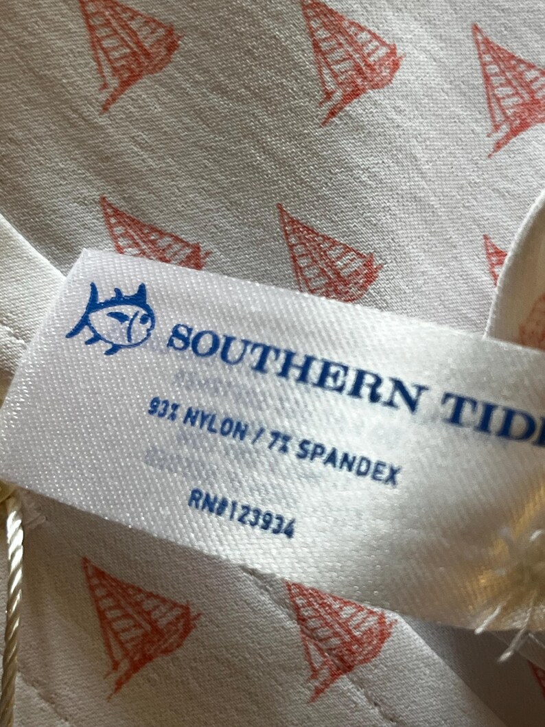 May include: Close-up of a white fabric with a repeating pattern of red sailboat illustrations. A white tag with blue text reads "SOUTHERN TIDE" and "93% NYLON / 7% SPANDEX". The tag also features a fish logo.
