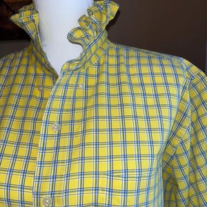 May include: A long-sleeved button-down shirt in a bright yellow, blue, and white plaid pattern. The shirt features a high, ruffled collar and a front button placket with light-colored buttons. A small embroidered logo is visible on the lower front.