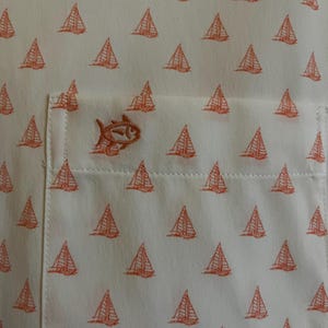 May include: Off-white button-up shirt with a repeating pattern of orange sailboats. The shirt features a chest pocket with an embroidered orange fish logo. The fabric has a subtle texture, and the overall design is nautical.