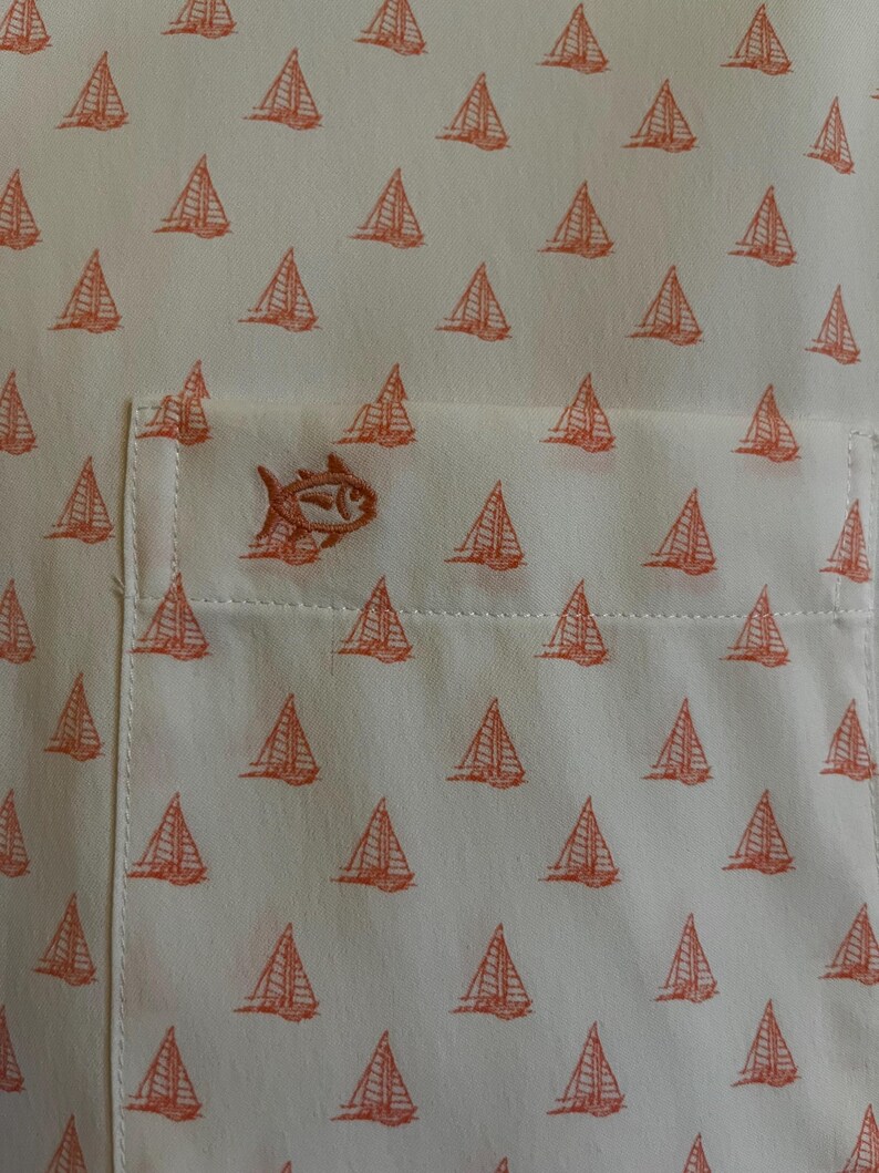 May include: Off-white button-up shirt with a repeating pattern of orange sailboats. The shirt features a chest pocket with an embroidered orange fish logo. The fabric has a subtle texture, and the overall design is nautical.