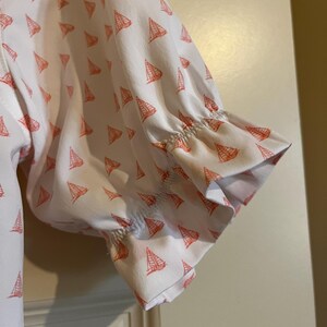 May include: White short-sleeved shirt with a pattern of small orange sailboats. The sleeves are gathered at the elbow and have a ruffled edge. The shirt is made of a lightweight fabric.