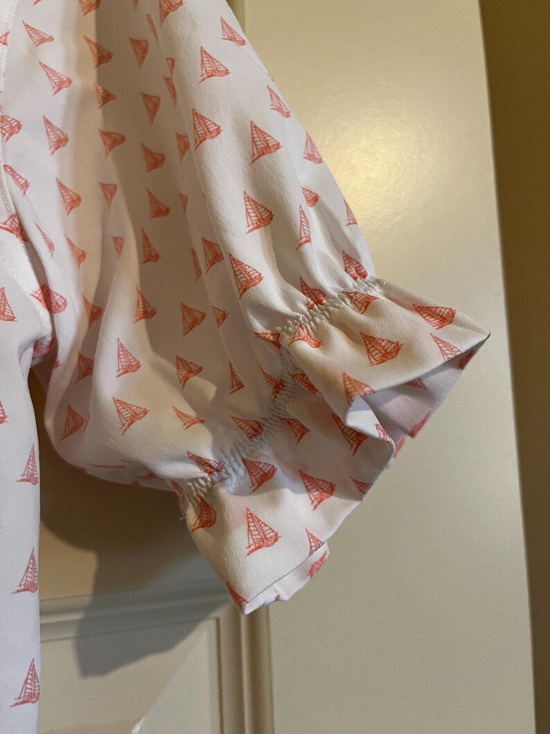 May include: White short-sleeved shirt with a pattern of small orange sailboats. The sleeves are gathered at the elbow and have a ruffled edge. The shirt is made of a lightweight fabric.