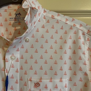 May include: White button-down shirt with a repeating pattern of small orange sailboats. The shirt has a chest pocket with a small fish logo and the brand name "Southern Tide". The shirt is XL size.