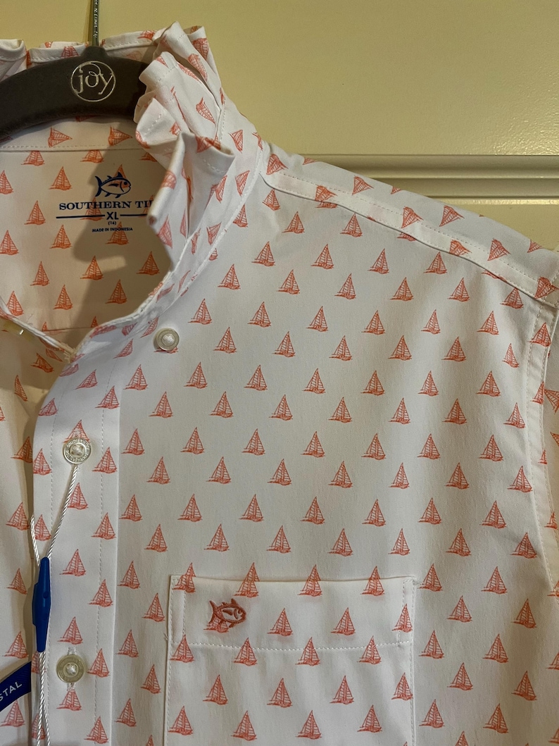 May include: White button-down shirt with a repeating pattern of small orange sailboats. The shirt has a chest pocket with a small fish logo and the brand name "Southern Tide". The shirt is XL size.