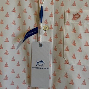 May include: Off-white button-down shirt with a pattern of small orange sailboats. The shirt has a chest pocket with an embroidered fish and a tag that reads "SOUTHERN TIDE". A blue tag reads "INTERCOASTAL".