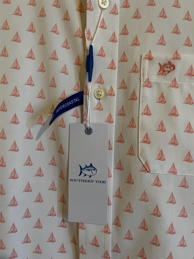 May include: Off-white button-down shirt with a pattern of small orange sailboats. The shirt has a chest pocket with an embroidered fish and a tag that reads "SOUTHERN TIDE". A blue tag reads "INTERCOASTAL".