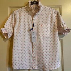 May include: White short-sleeve shirt with a pattern of small pink sailboats. The shirt has a ruffled collar and cuffs, a chest pocket, and a button-down front. A tag with the brand name is attached.