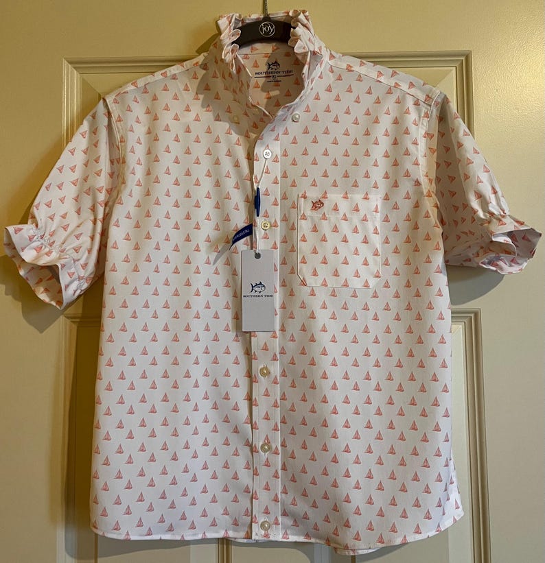 May include: White short-sleeve shirt with a pattern of small pink sailboats. The shirt has a ruffled collar and cuffs, a chest pocket, and a button-down front. A tag with the brand name is attached.