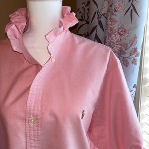 May include: A light pink button-down shirt featuring a ruffled collar and short sleeves with ruffled cuffs. The shirt has a front button placket and a small embroidered logo on the left chest area, displayed on a white mannequin torso.