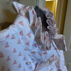 May include: White button-down shirt with a repeating pattern of red sailboats. The shirt features a ruffled collar and cuffs. The fabric is lightweight and appears to be cotton or a cotton blend. The shirt is hanging on a hanger.