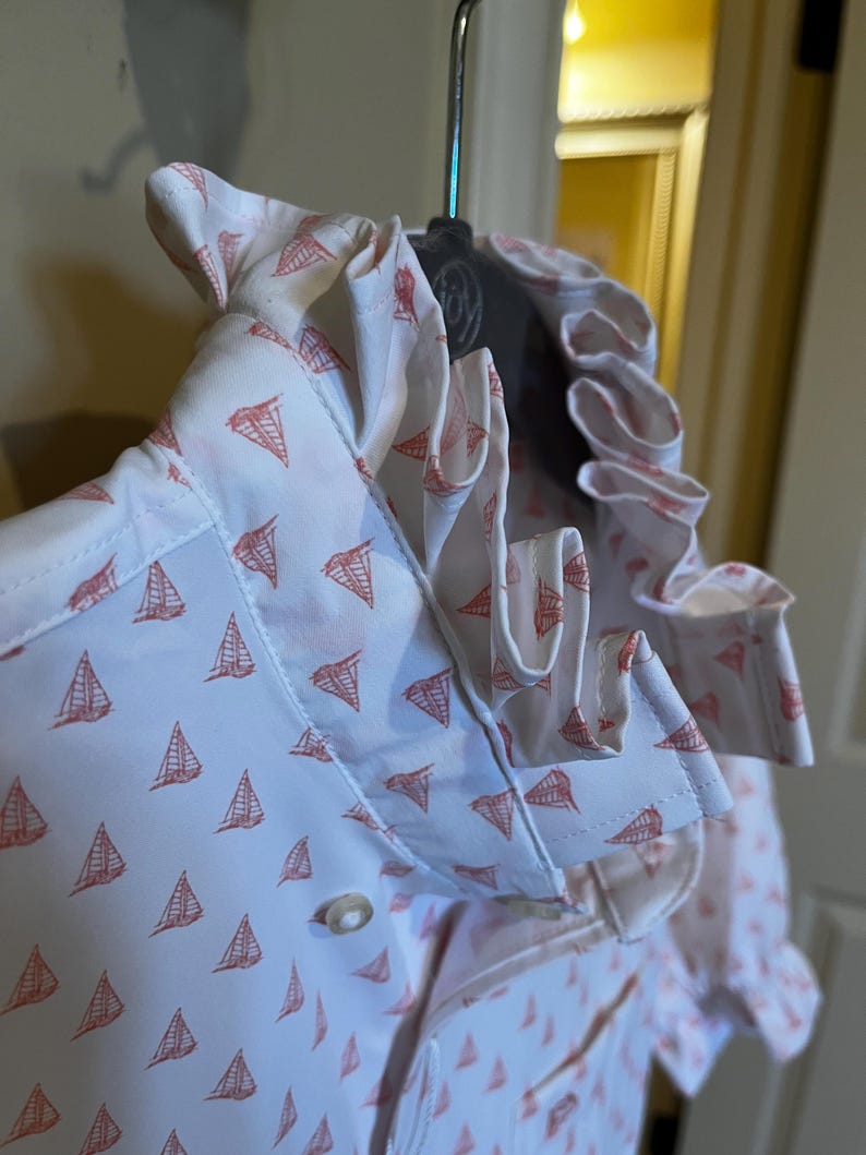May include: White button-down shirt with a repeating pattern of red sailboats. The shirt features a ruffled collar and cuffs. The fabric is lightweight and appears to be cotton or a cotton blend. The shirt is hanging on a hanger.