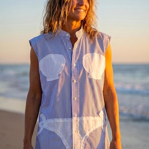 May include: A sleeveless blue and white pinstriped button-down shirt featuring two white shell-shaped appliqués on the chest and a white bikini-style graphic appliqué across the lower front section, complete with a mandarin collar and front buttons.