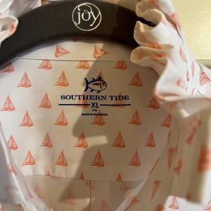 May include: White button-down shirt with a repeating orange sailboat pattern. The shirt features the "Southern Tide" logo and is size XL (14). The shirt is hanging on a dark gray hanger with the word "joy" embroidered on it.