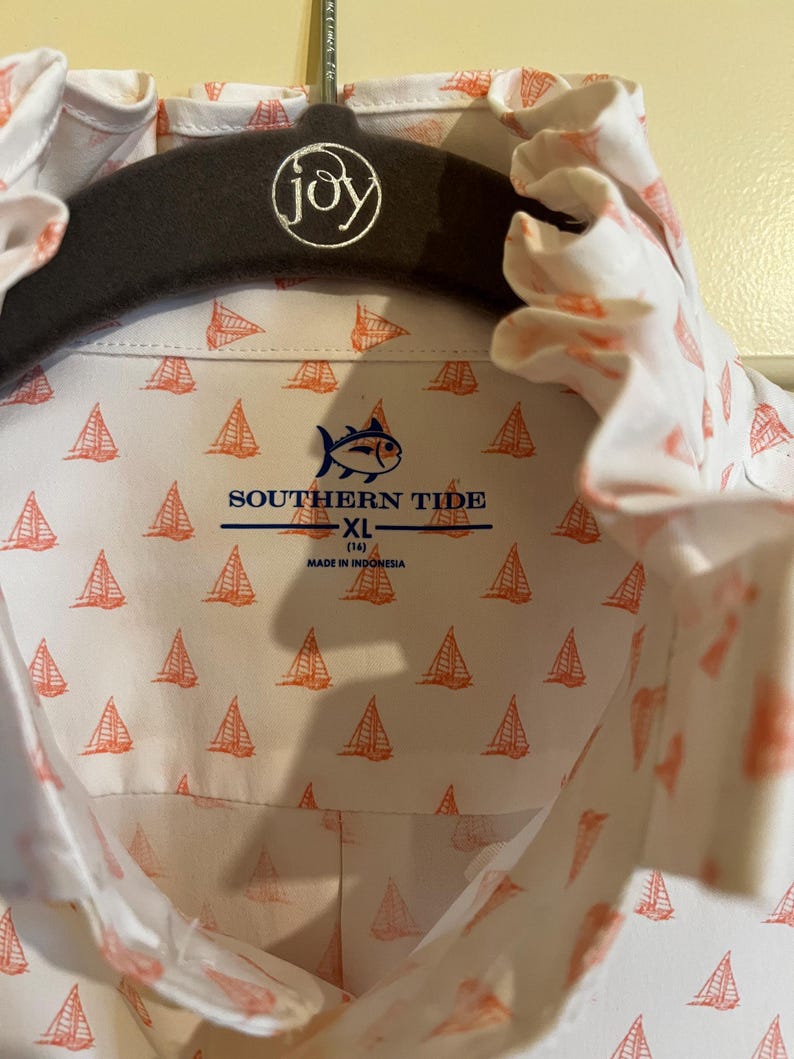 May include: White button-down shirt with a repeating orange sailboat pattern. The shirt features the "Southern Tide" logo and is size XL (14). The shirt is hanging on a dark gray hanger with the word "joy" embroidered on it.