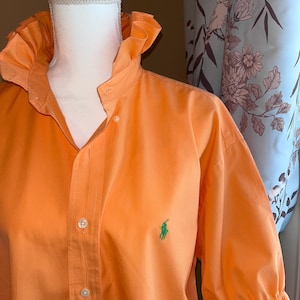 May include: A bright orange button-down shirt featuring a ruffled collar and ruffled sleeve cuffs. The shirt has a front button placket with white buttons and a small green embroidered logo on the left chest area, displayed on a white mannequin.