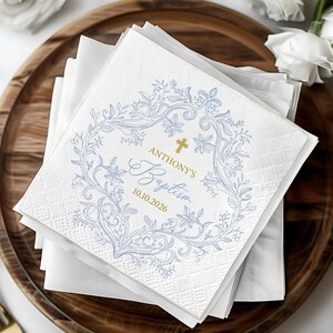 May include: A stack of white square paper napkins featuring a light blue floral wreath design. In the center, a gold cross sits above the text "ANTHONY'S Baptism" and the date "10.10.2026" printed in a combination of gold and blue script fonts.