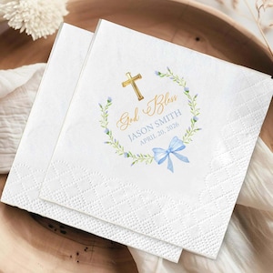 May include: Two white paper napkins with a diamond pattern. One napkin features a gold cross, the words "God Bless", a name, date, and a blue bow within a floral wreath. The other napkin is plain.