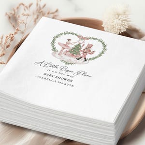 May include: A stack of white paper napkins featuring a watercolor illustration of a ballerina and a nutcracker beside a Christmas tree, framed by a green wreath. The text reads: A Little Sugar Plum is on her way BABY SHOWER ISABELLA MARTIN.