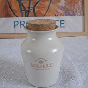 May include: A small, off-white ceramic jar with a tapered neck and a round cork stopper. The front of the jar features the gold-colored text "MAILLE" with a small emblem above it and smaller text below, all centered on the smooth, glossy surface.