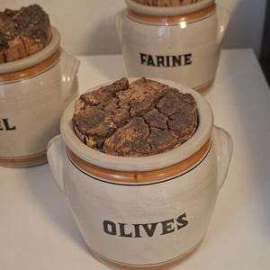 May include: Three off-white ceramic storage jars with two side handles and brown horizontal bands. Each jar features a rough, textured, dark brown cork lid. The front of the jars are labeled with black text reading OLIVES, FARINE, and a partial label.