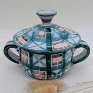 May include: A ceramic sugar bowl with a lid, featuring a teal and white checkered pattern. The bowl has two handles and a small knob on the lid. The design includes brown accents. Dried ornamental grass is in the foreground.