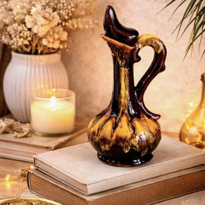 Peut inclure: Pichet décoratif en céramique avec un glaçage marron foncé et jaune. Le pichet a une poignée incurvée et un bec avec un couvercle. Il est placé sur deux livres. L'arrière-plan comprend un vase avec des fleurs séchées et une bougie allumée.