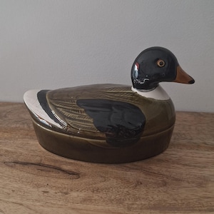 Peut inclure: Un récipient en céramique en forme de canard avec un couvercle et une finition brillante. Le canard présente une tête vert foncé, un corps brun et noir avec des détails d'ailes, un collier blanc et un bec beige. La base est un bol ovale lisse.