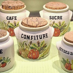 Può includere: Una collezione di barattoli da cucina in ceramica con coperchi in sughero. Ogni barattolo è bianco e presenta una parola diversa in nero: CAFE, SUCRE, CONFITURE, THE e HERBES DE PROVENCE. I barattoli sono decorati con illustrazioni colorate di verdure ed erbe.